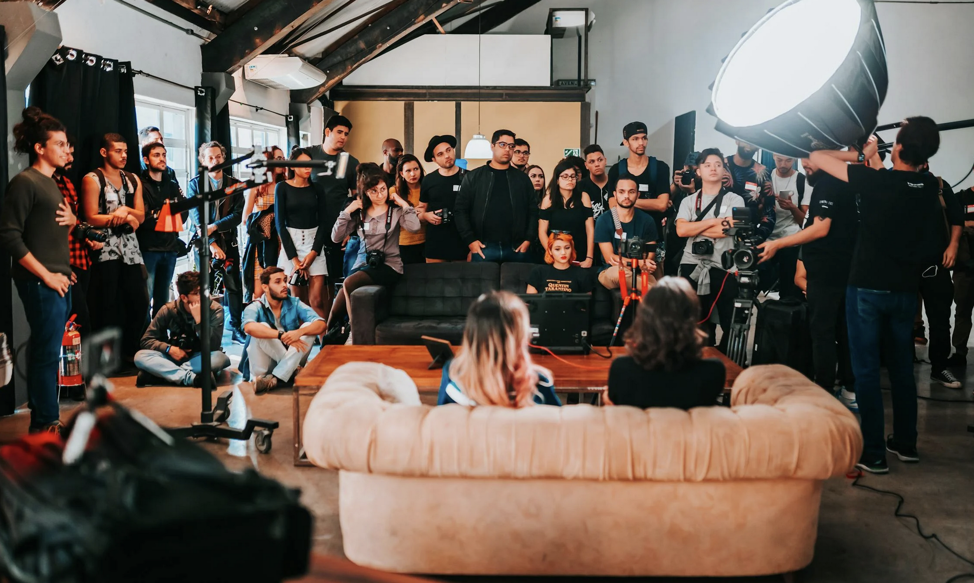 dwie osoby odwrócone tyłem, siedzące na kanapie w studiu, otoczone pracownikami mediów i widownią