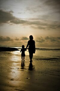 widok od tyłu na matkę trzymającą dziecko za rękę, spacerują po plaży w kierunku zachodzącego słońca
